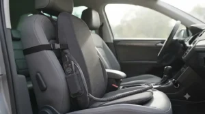 The charcoal grey fabric massage cushion (from image_2.png) is installed on the grey leather driver's seat of a modern car. It is secured with the adjustable strap, and the coiled power cable is plugged into the 12V socket (cigarette lighter) near the gear stick.