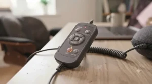 A detailed close-up photograph of the wired remote control for the massage cushion, resting on a wooden desk. The remote is black with clearly labelled, tactile buttons for Power, Mode, Heat, and Intensity (+/-), and a small red indicator light.